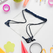A clear drawstring bag with navy blue accents is centered on a white desk with various office supplies scattered around it.
