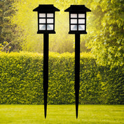 Two black solar garden lights with white paneled tops stand in a vibrant green lawn with hedges and trees.