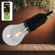 A black and clear portable LED tent light with an orange filament lies diagonally on a wooden surface, with its green and