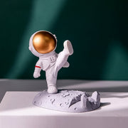 Astronaut Phone Holder (kicking leg design) on a wooden desk, holding a smartphone.