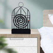 A black iron birdcage-style holder with a burning mosquito coil sits on a light wood nightstand next to a bed.