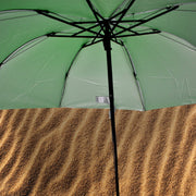 The underside of an open green umbrella reveals its black frame and silver lining, above a textured brown surface.
