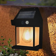 A black solar wall lamp with a clear bulb glowing warmly is mounted on a textured brown wall above green plants.