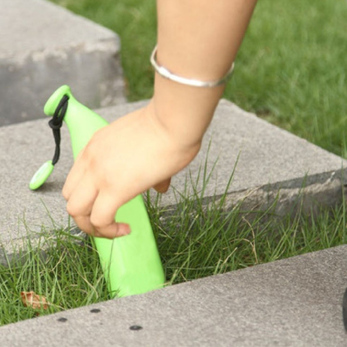 A hand reaches for a green banana-shaped umbrella case resting in green grass between concrete slabs.