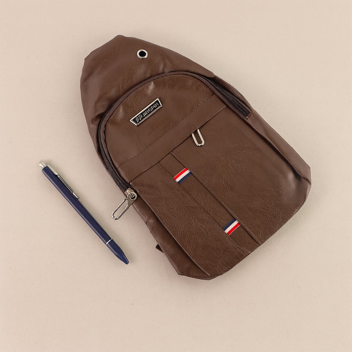 A brown leather sling bag lies flat on a light brown surface with a blue pen next to it.
